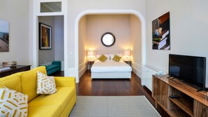 Un soggiorno moderno con un divano giallo, un letto bianco con due cuscini, uno specchio rotondo sopra il letto e un televisore su un supporto in legno.