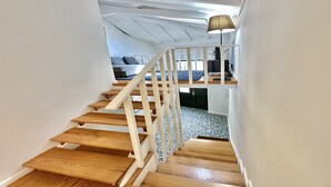 Una scala in legno con ringhiere bianche conduce a una camera da letto soppalcata con un letto e una lampada.