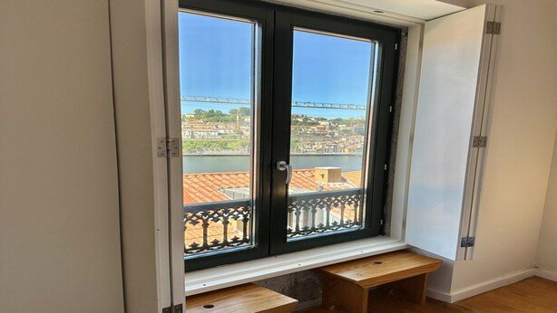 Una stanza con una grande finestra che si affaccia su un panorama urbano e un ponte. Sotto la finestra, una panca in legno.