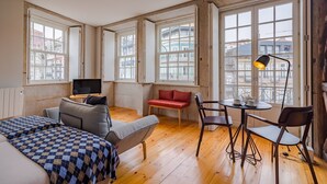 A spacious room with a large bed, a sofa, a red bench, and a dining table with chairs. There are multiple windows providing a view of buildings outside. - Pretty apartment with modern comforts (Oporto)