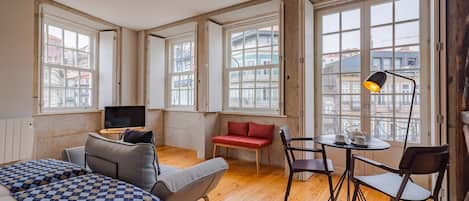 A spacious room with a large bed, a sofa, a red bench, and a dining table with chairs. There are multiple windows providing a view of buildings outside.