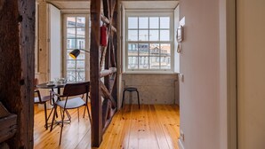 A room with wooden floors, a dining table with chairs, a small stool, and a window with a view of buildings. - Pretty apartment with modern comforts (Oporto)