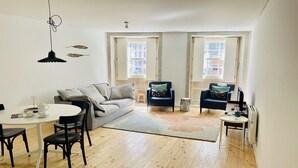 A modern living room with a sofa, two armchairs, a dining table with chairs, and a small side table. There are two windows with open shutters, a hanging light fixture, and a radiator.