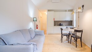 A compact living space with a sofa, a dining table with chairs, a kitchen area, and a ceiling-mounted air conditioner.