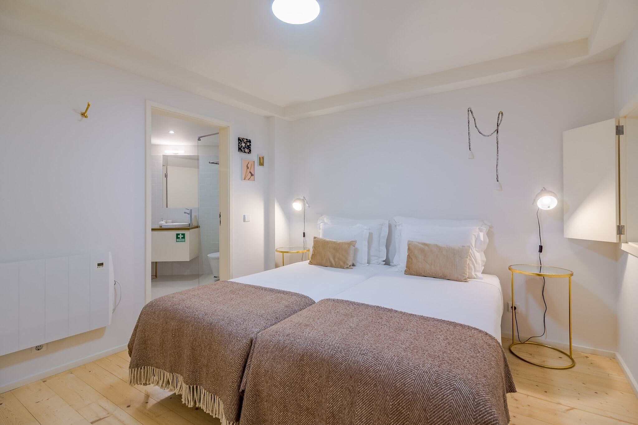 Quarto com cama de casal, duas mesas de cabeceira, um abajur, um radiador e uma pequena mesa redonda com uma luminária.