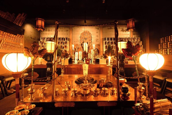 Un montaje de comedor tradicional con una mesa central, múltiples velas y un fondo de estanterías con diversos objetos.
