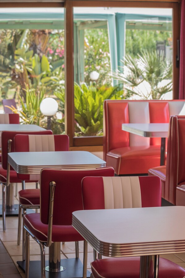 A restaurant with red booths and white tables, large windows with greenery outside.