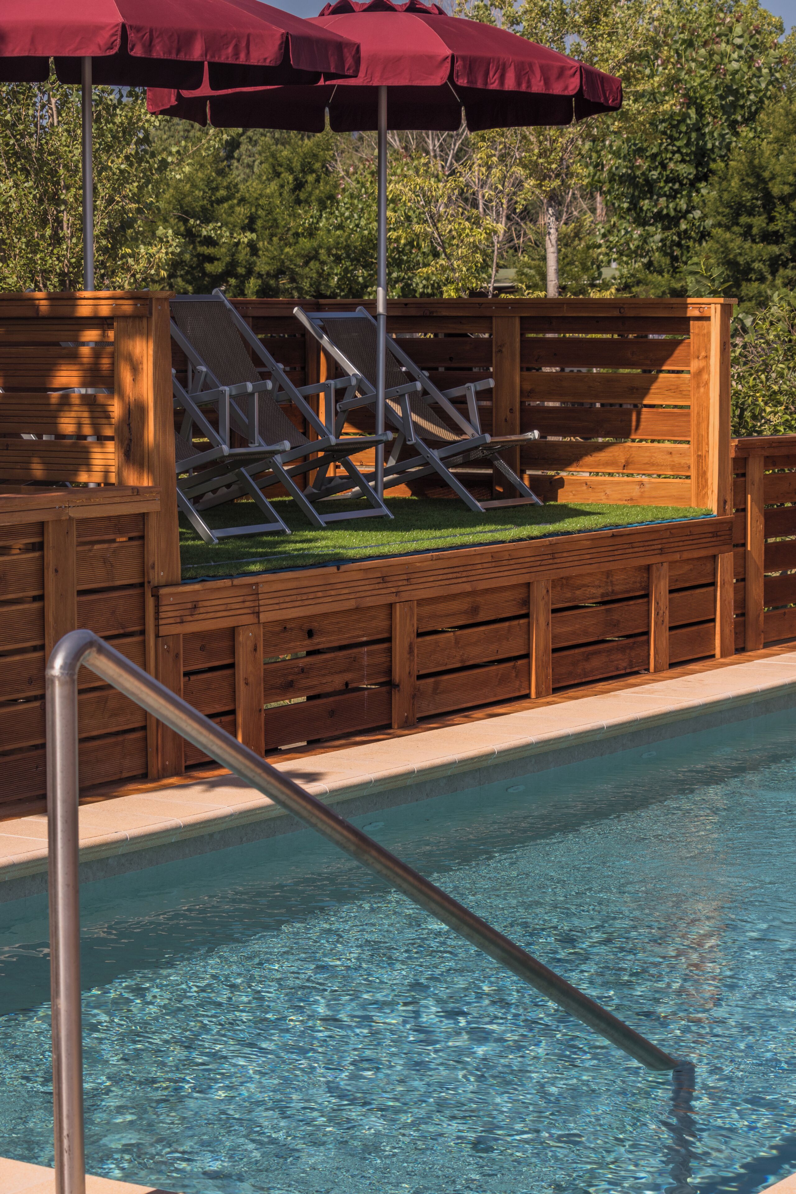 Una piscina con muelle de madera, una escalera metálica y una sombrilla roja.