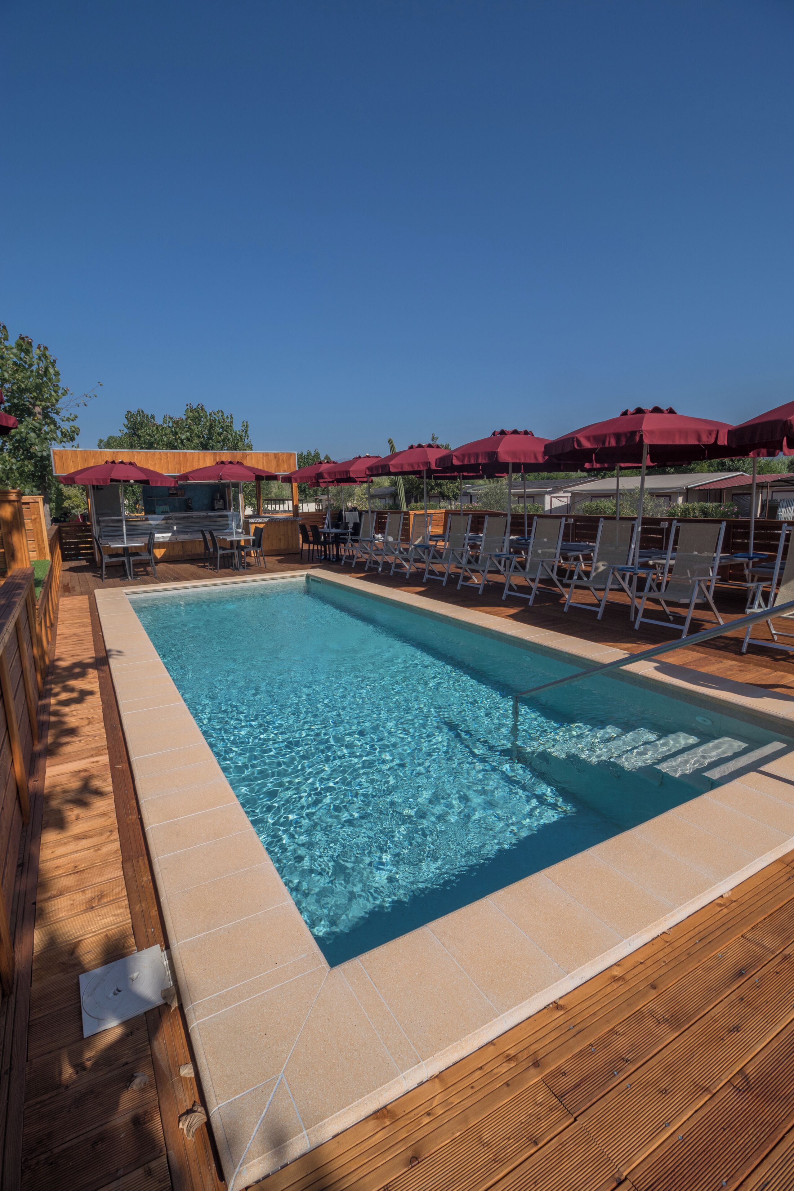 Piscina rectangular con agua azul cristalina, rodeada de un deck de madera y sillones de descanso.