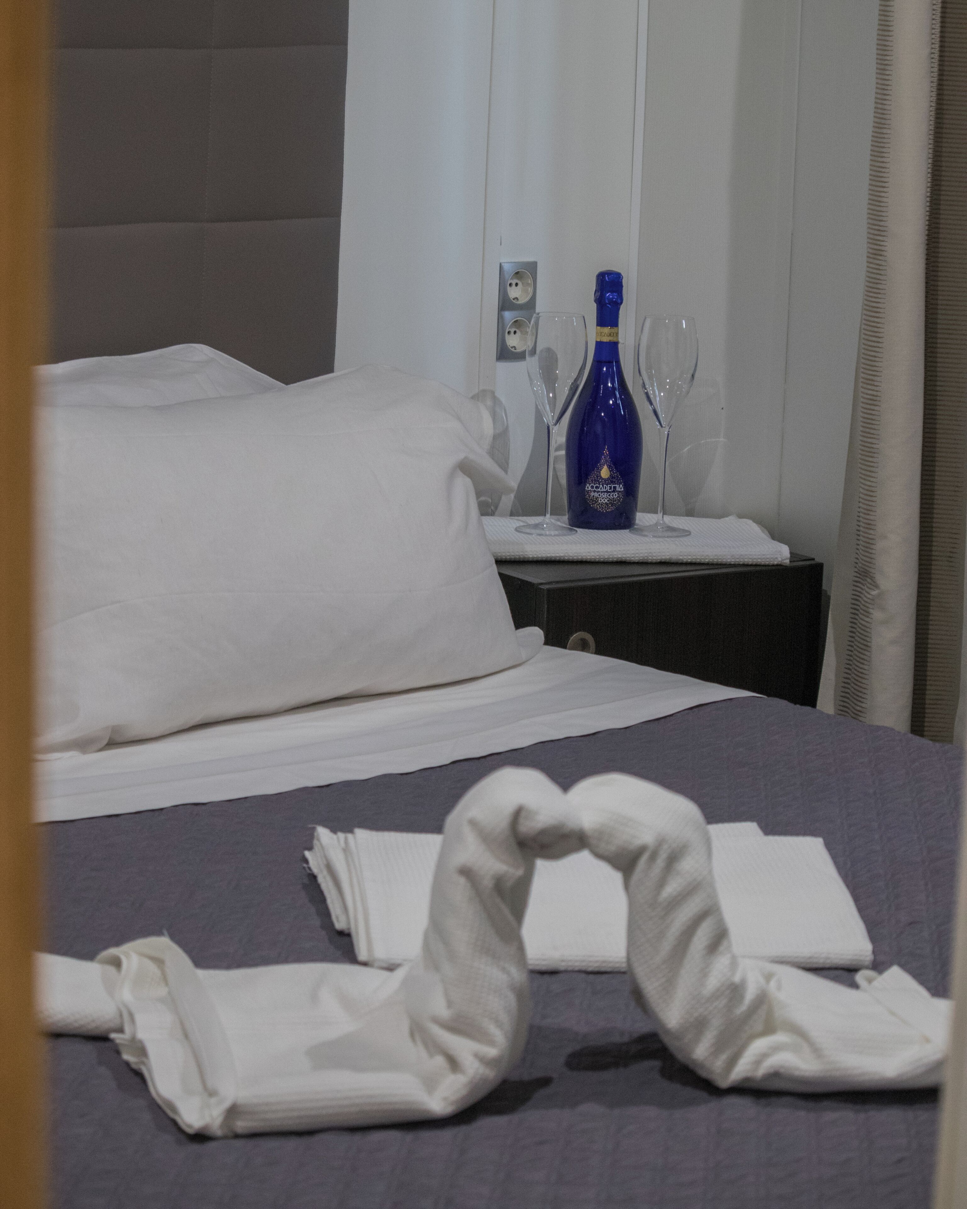 A neatly made bed with white linens, a bottle of blue liquid, and two wine glasses on a bedside table.