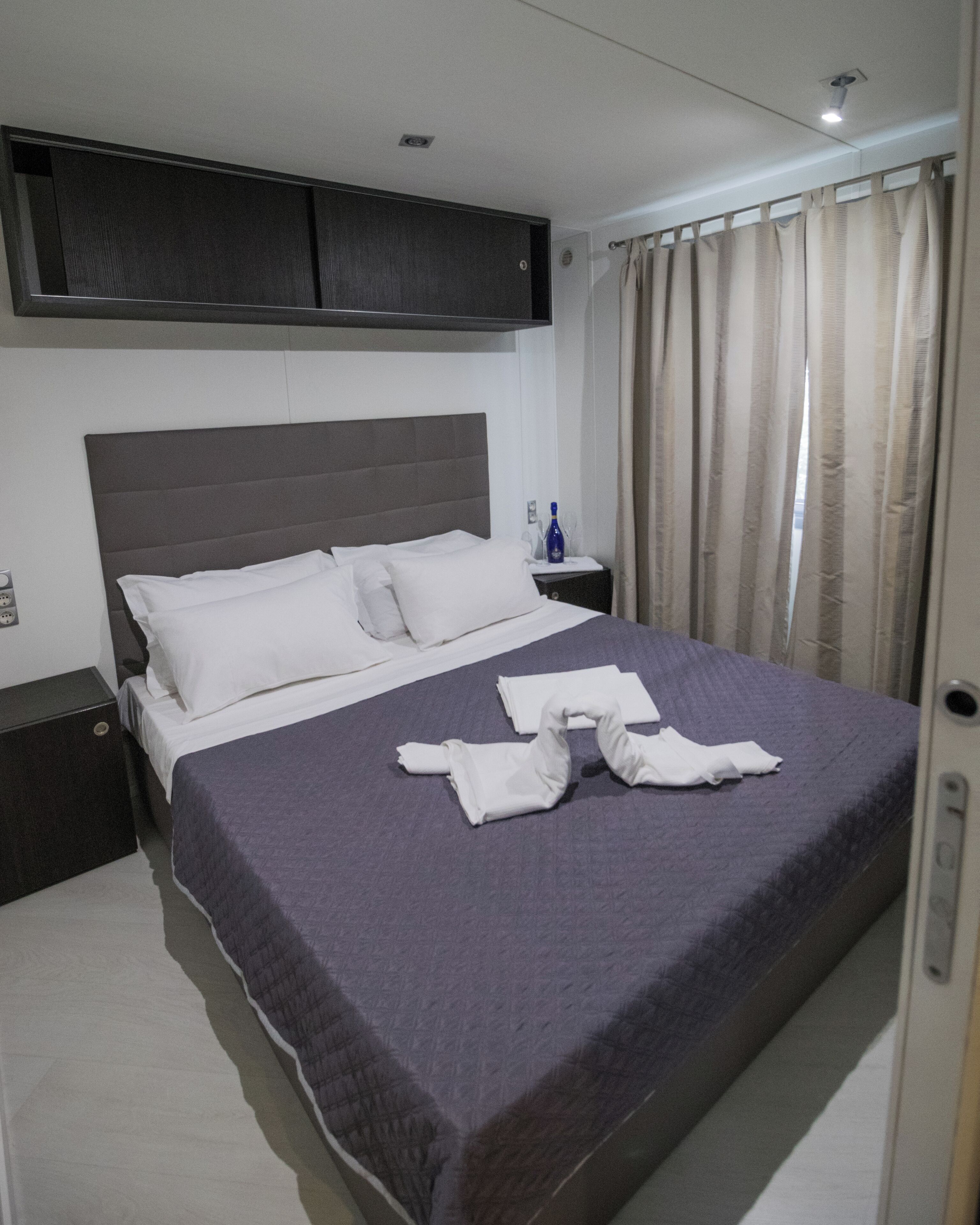 A neatly made bed with a gray headboard, a bedside table with a bottle, and a curtain on the window.