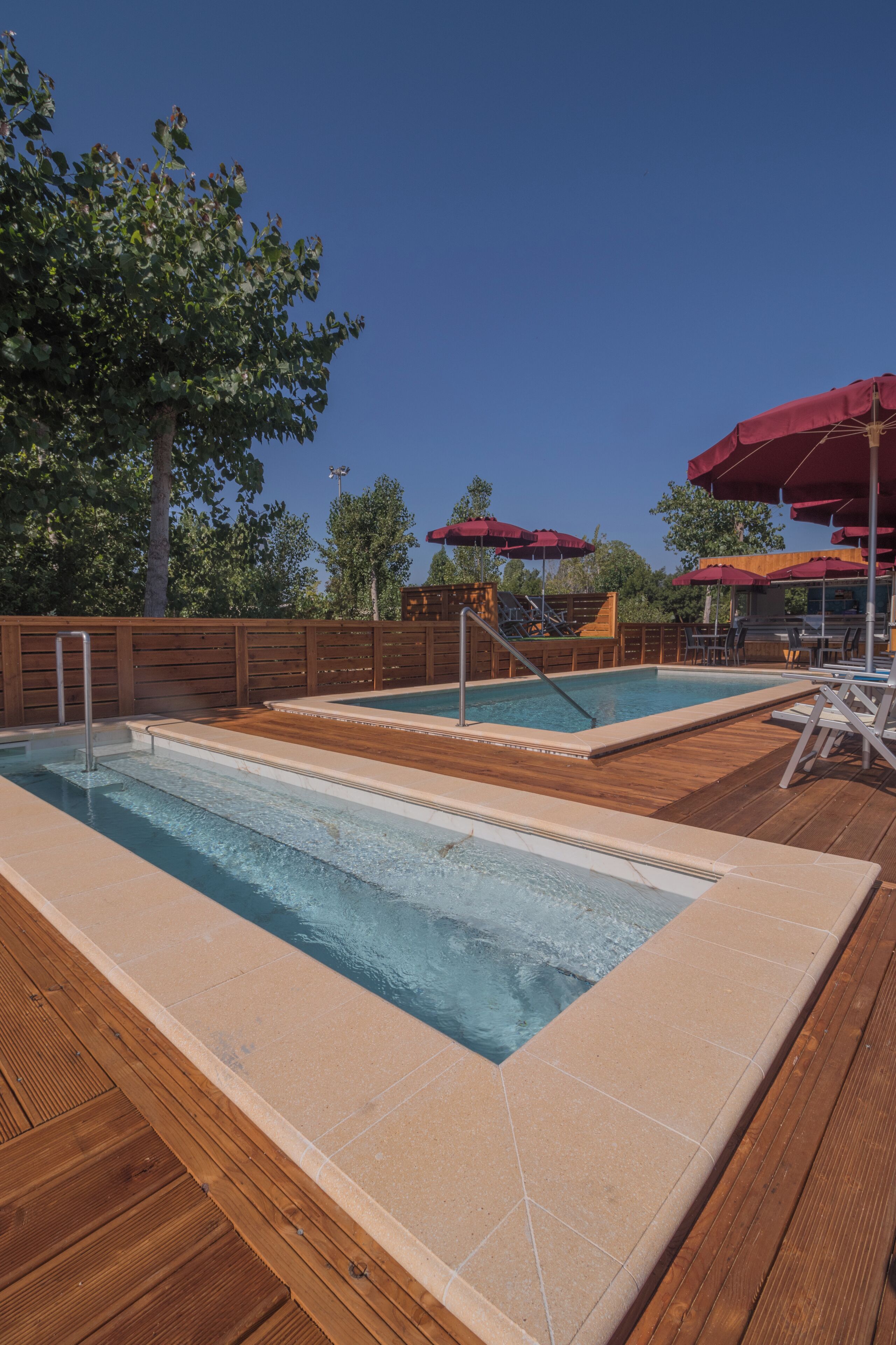 Zona de piscina con dos piscinas, cubierta de madera y sombrillas rojas.