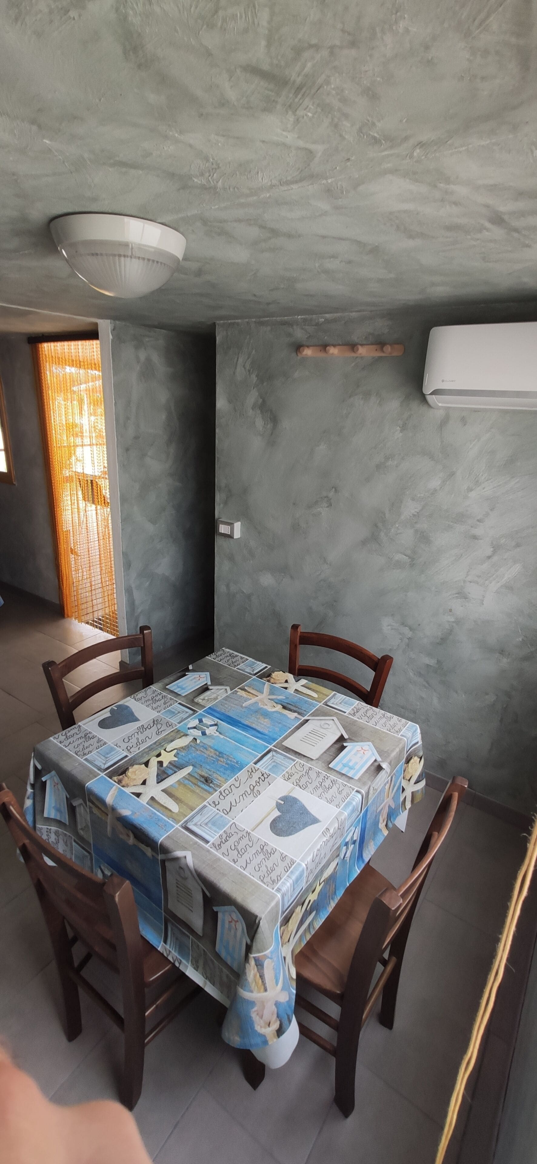 A dining area with a table covered in a patterned tablecloth, four wooden chairs, a ceiling light fixture, and a wall-mounted air conditioning unit.