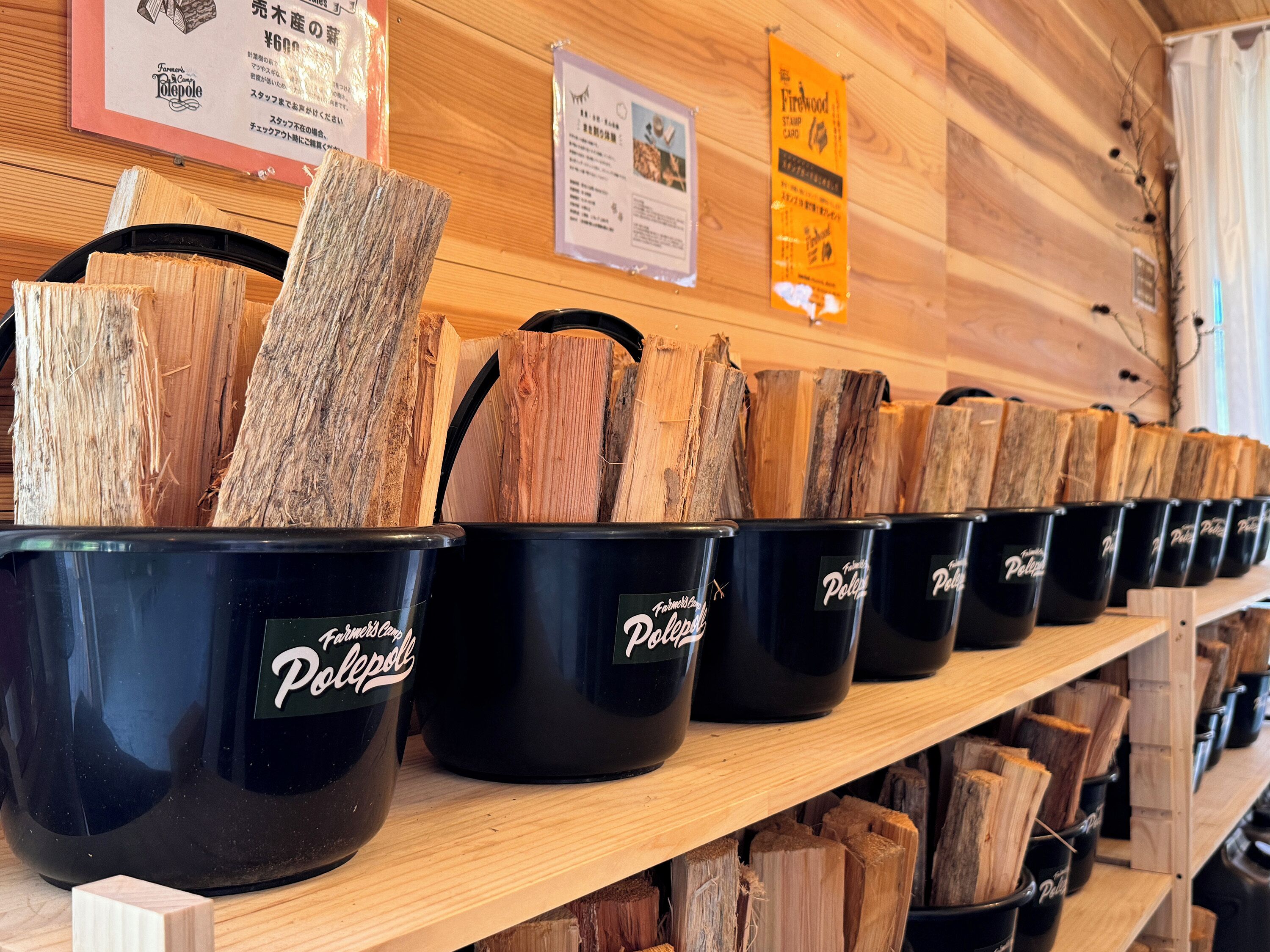 A row of black buckets labeled "Farmers' Country Polepole" on a wooden shelf, with wood logs inside.