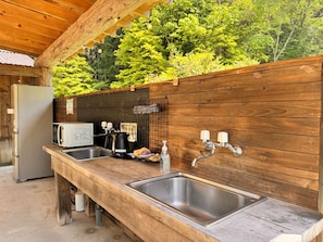 A rustic outdoor kitchen with wooden counters, a sink, a kettle, and a refrigerator.
