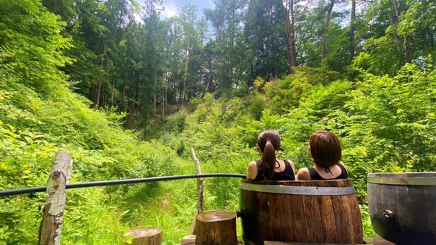 Deux personnes sont assises dans une baignoire en bois sur une terrasse en bois, entourées d’une végétation luxuriante.