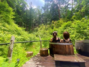Two people sitting in a wooden tub on a wooden deck surrounded by lush greenery. - Polepole Village (Urugi)