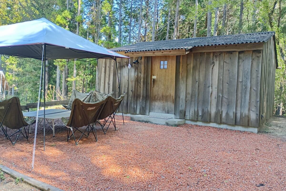 Une cabane en bois rustique, dotée d’une porte et d’un petit porche, entourée d’une zone recouverte de gravier, avec une table et des chaises placées sous une tonnelle.