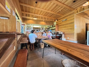 A wooden interior with tables and benches, a counter with a person behind it, and shelves with various items. - Polepole Village (Urugi)