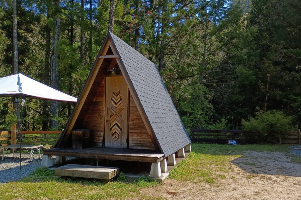 Une structure en bois en forme de « A », avec une porte, un banc et une table placés sous une tente.