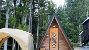 Une cabane en bois avec un toit triangulaire et une porte ornée d’un motif en chevrons. Une tente est installée à proximité.