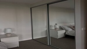 Quarto com porta de correr de vidro, mesa de cabeceira branca, abajur e cama com travesseiros e cobertor.
