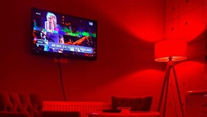 A room with a television mounted on the wall displaying a news channel, a table with chairs, and a floor lamp. - ÇANKAYA BOUTİQUE HOTEL (Ankara)