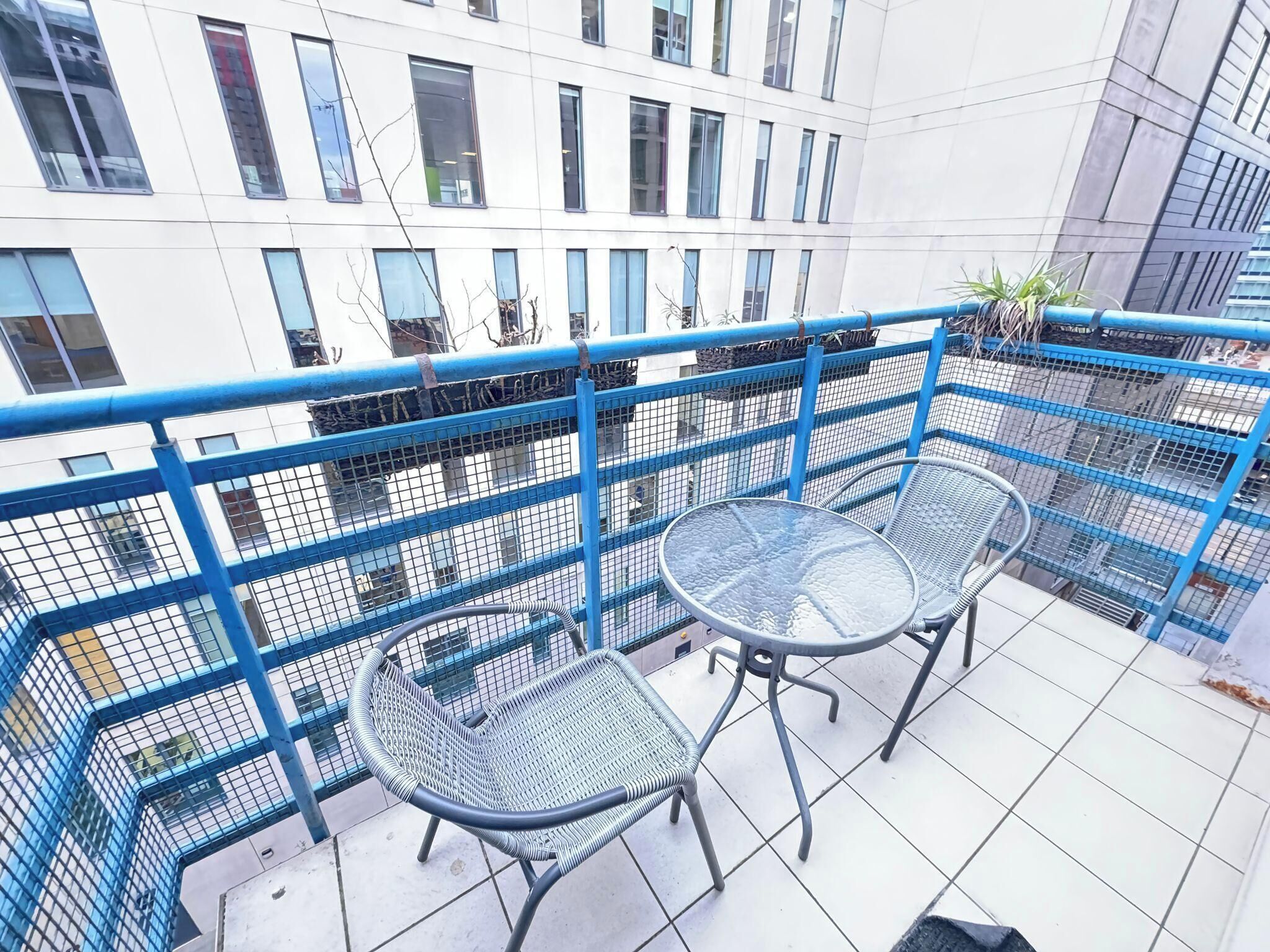 A balcony with two metal tables and chairs, a blue metal railing, and a view of a white building with multiple windows.