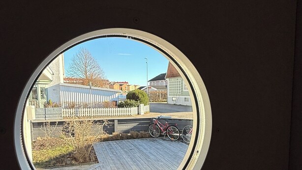 Et rundt vindue der viser udsigt til en træterrasse, et hvidt hegn og en bygning i baggrunden.