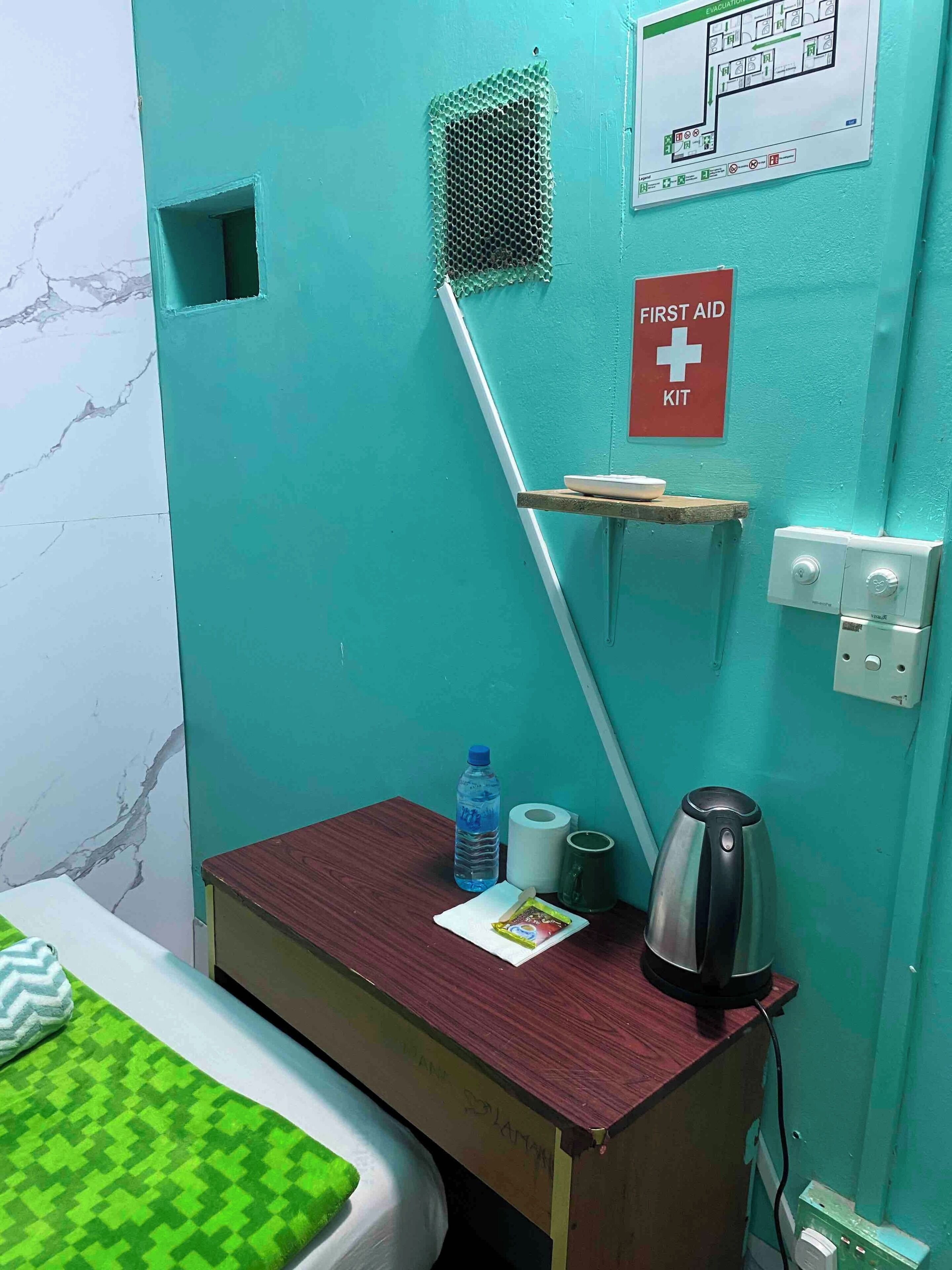 Een kamer in teal met een bed, een tafeltje met een waterfles, een waterkoker en een bordje met daarop een verbandtrommel.