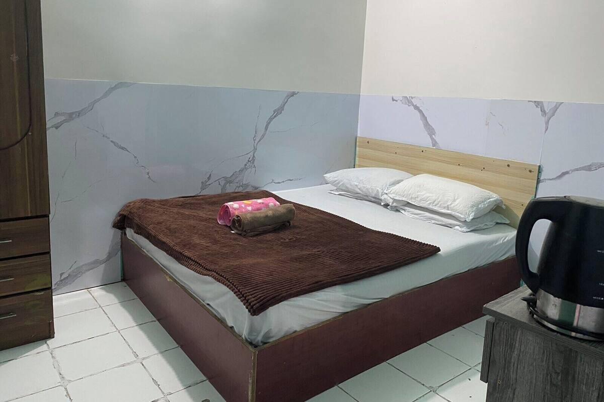 Une chambre à coucher avec un lit, des oreillers, une couverture, une armoire en bois et une bouilloire posée sur une table de chevet.