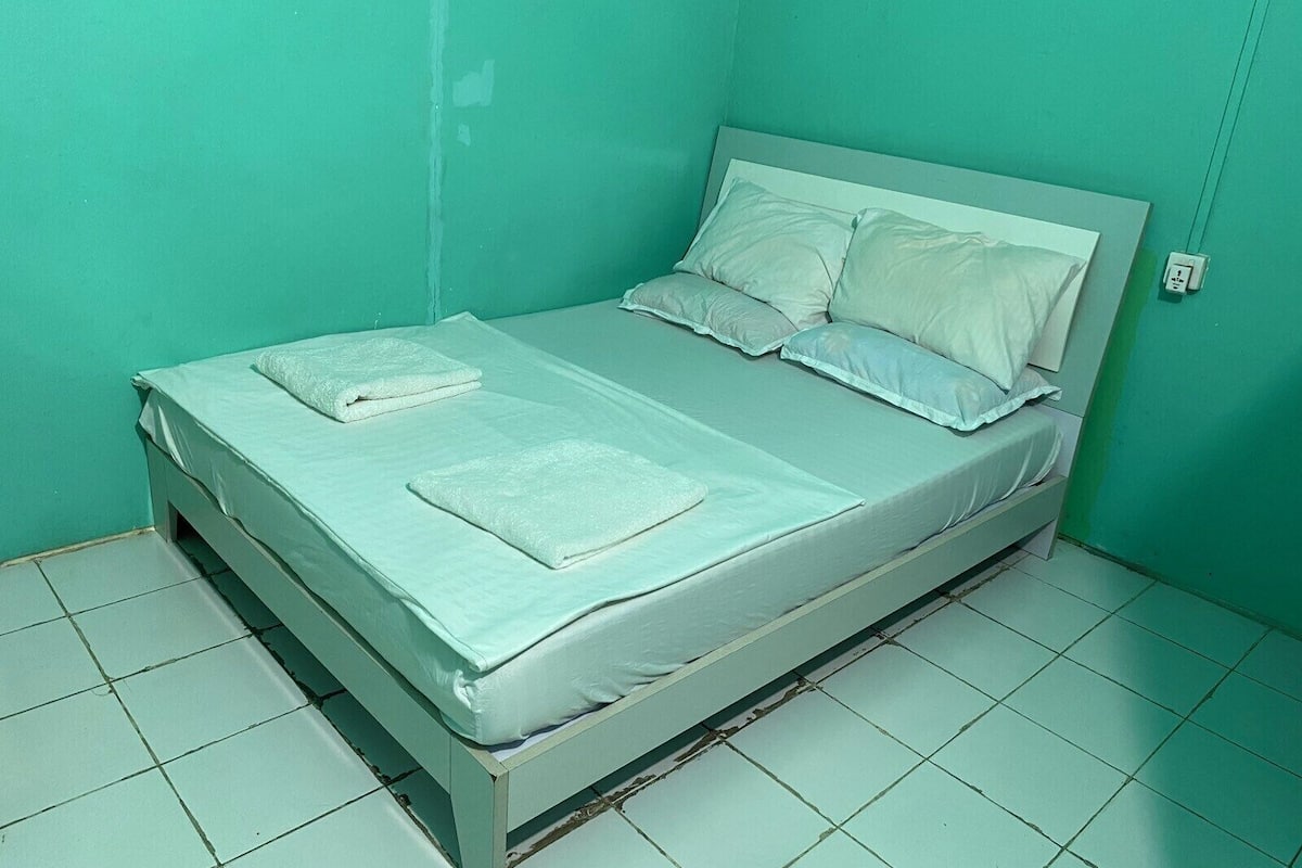 Une chambre avec un lit simple, un matelas blanc, deux oreillers et une couverture pliée.