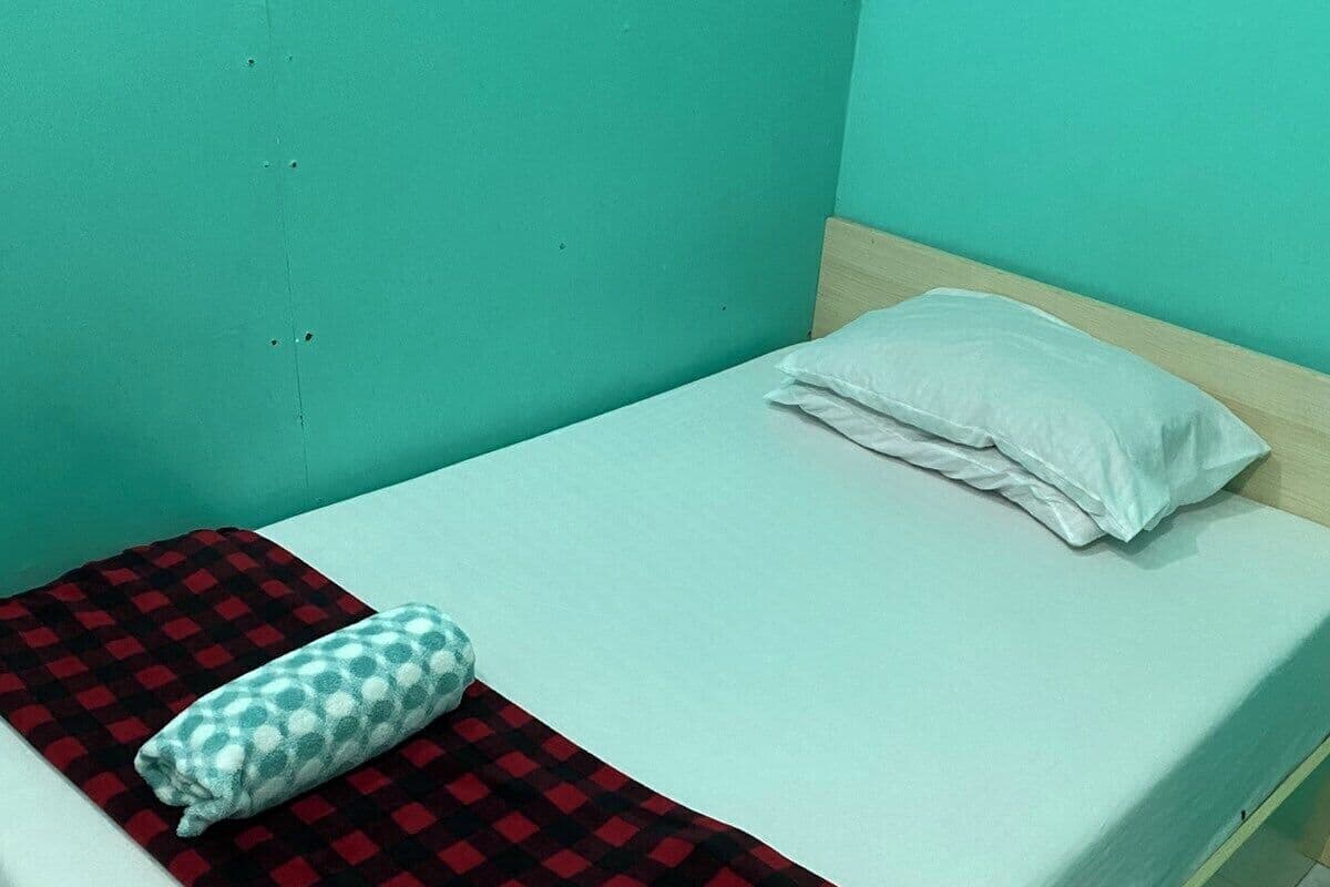 Un lit simple avec une couverture à carreaux rouges et noirs, un oreiller blanc et un autre oreiller à motifs.