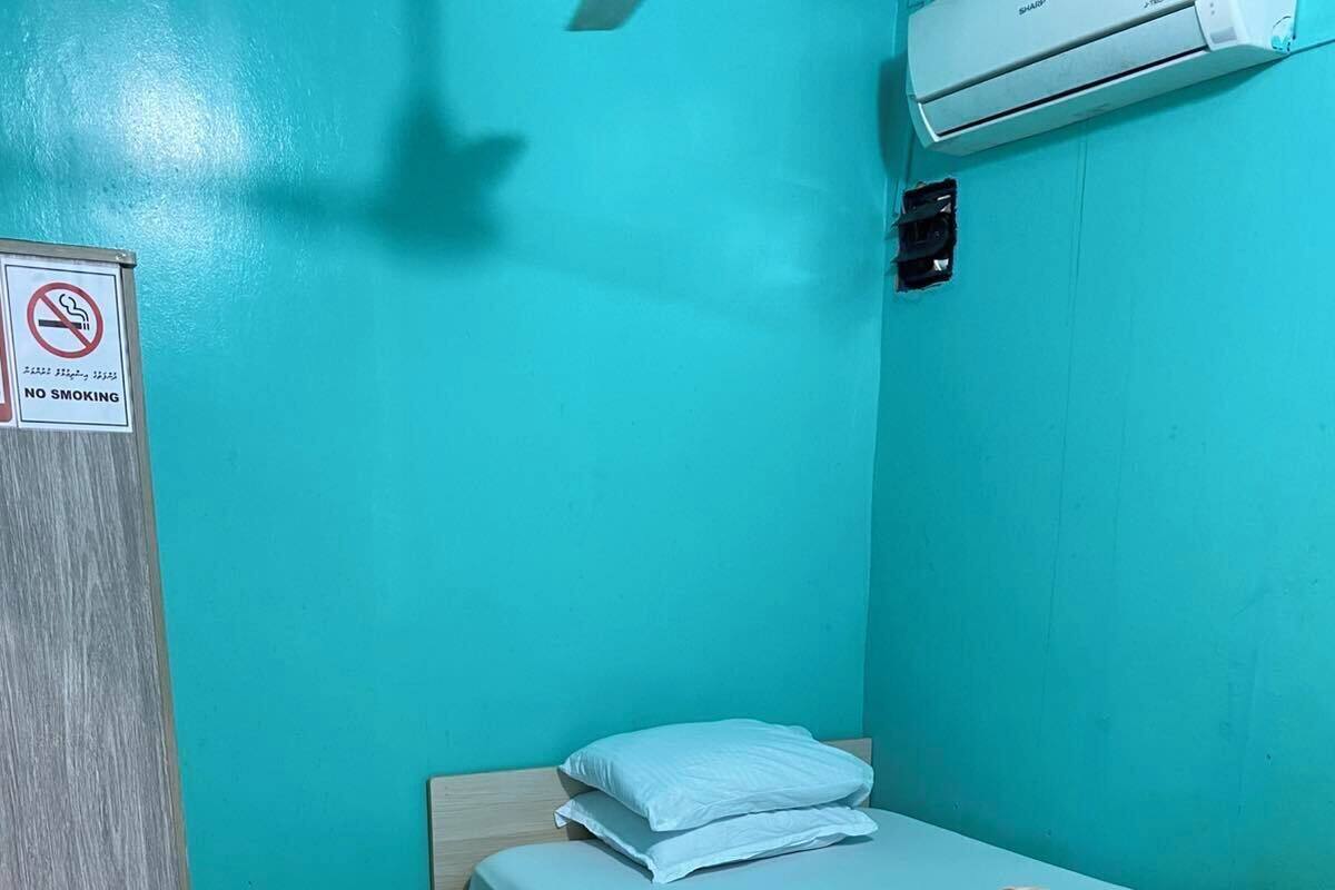 Une petite chambre avec un lit simple, un mur bleu, un sol à motifs, un panneau «interdiction de fumer» et un appareil de climatisation.