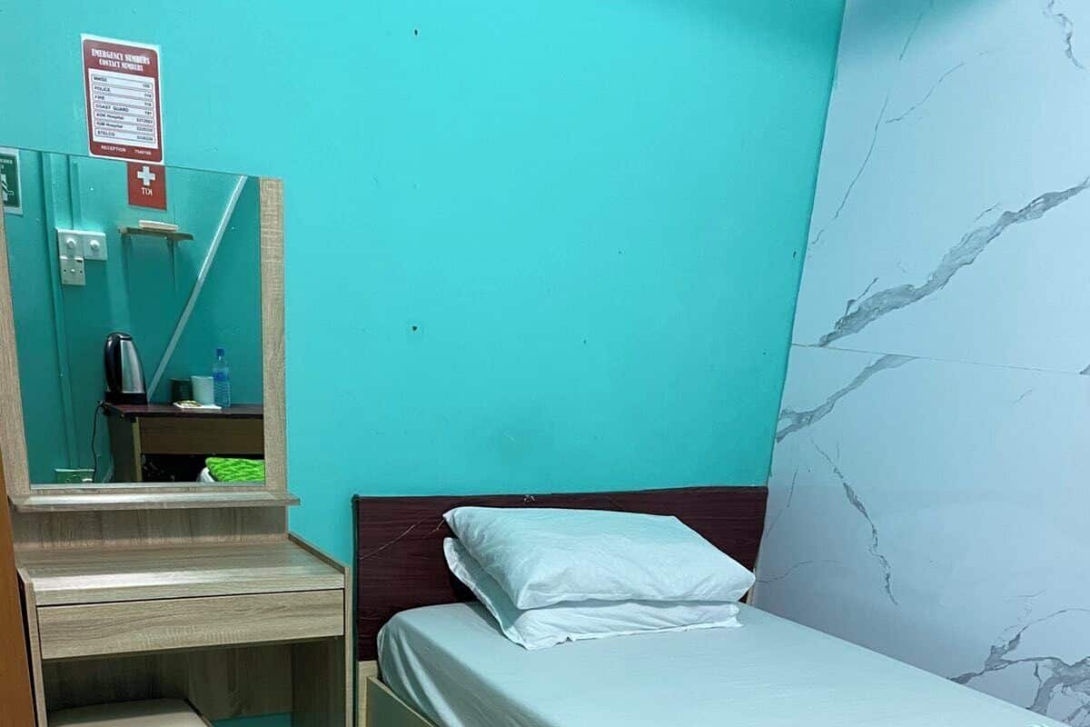 Une petite chambre avec un lit simple, un bureau en bois, une chaise, une couverture à motifs verts et une liste de numéros d’urgence affichée sur le mur.