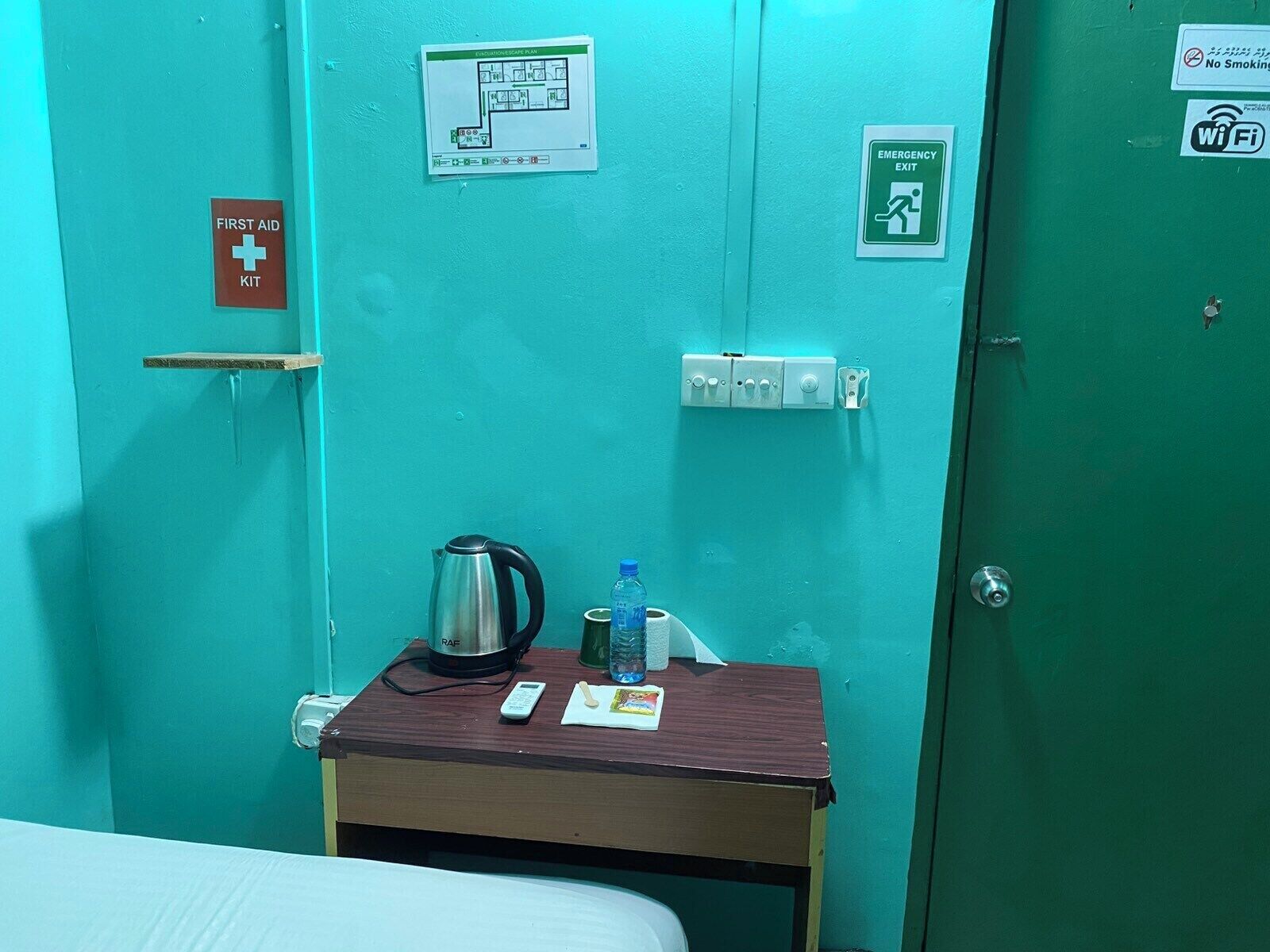 Une pièce aux murs de couleur turquoise, avec un lit, une petite table sur laquelle se trouvent une bouilloire, une bouteille d’eau et une télécommande. On y trouve également des panneaux indiquant l’emplacement d’une trousse de premiers secours, d’une sortie de secours et interdisant de fumer.