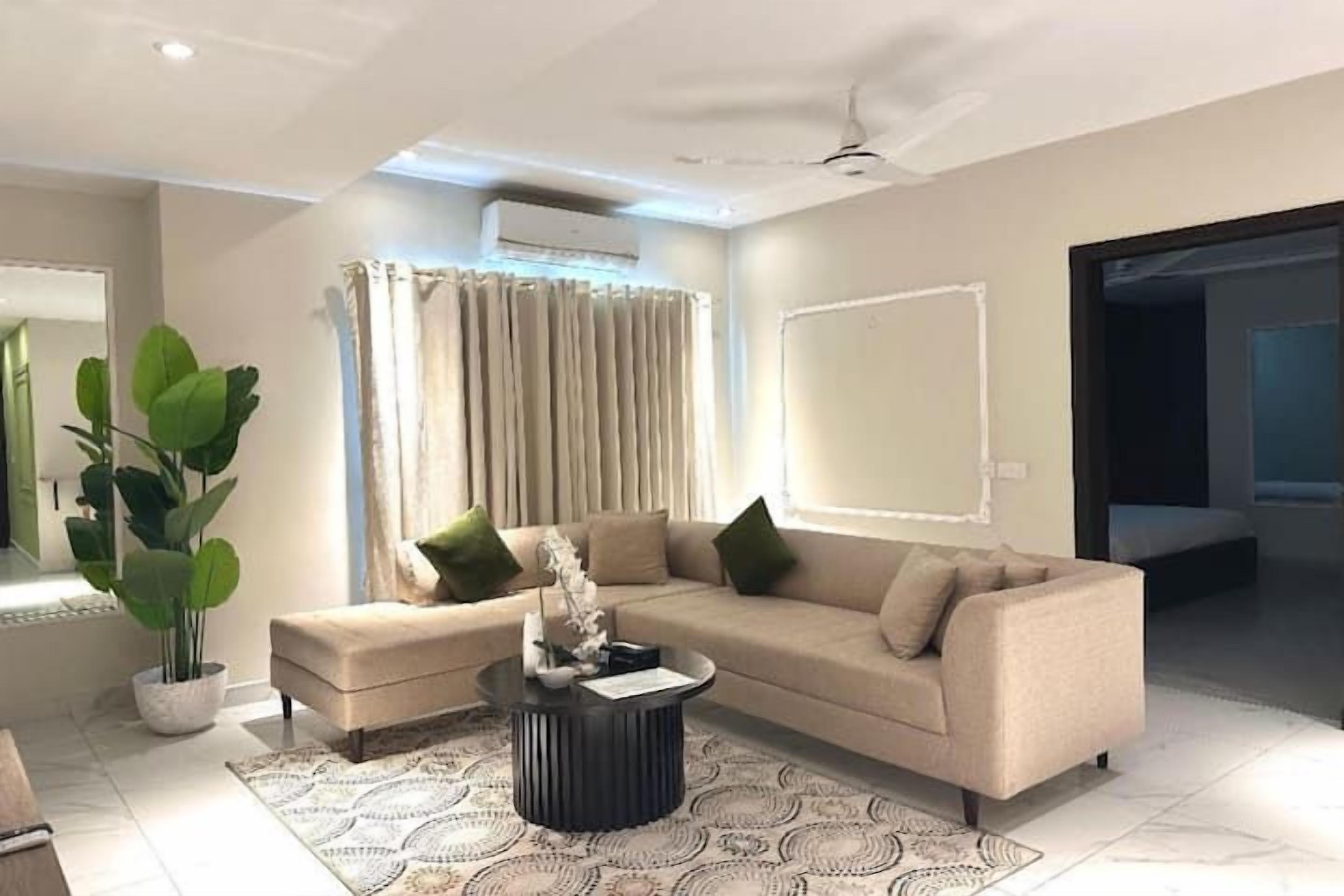 A modern living room with a sofa, ottoman, and coffee table. There is a potted plant and a large window with curtains.