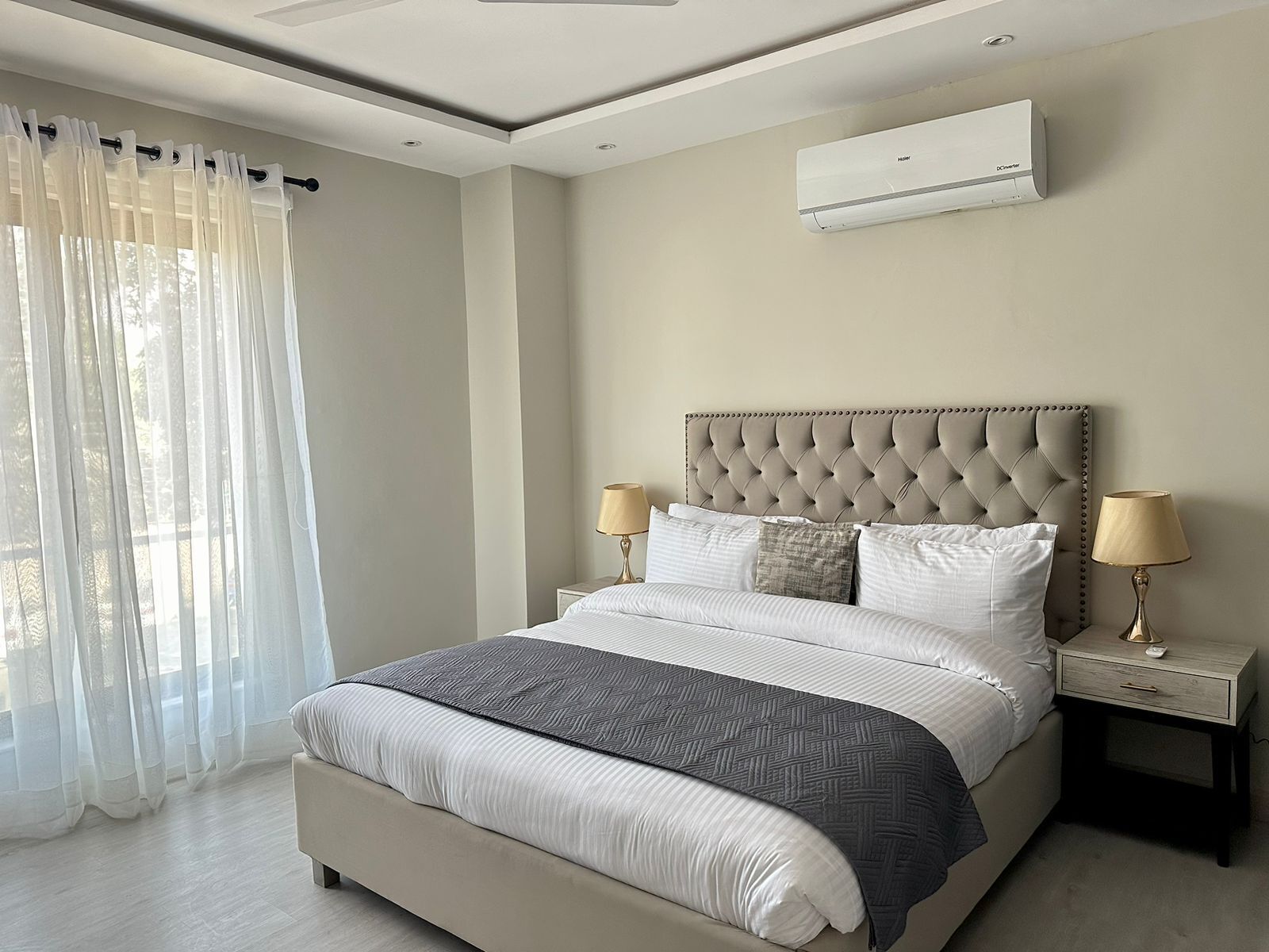 A modern bedroom with a tufted headboard, a large bed with multiple pillows, a bedside table with lamps, and an air conditioning unit on the wall.