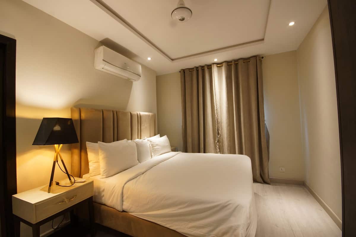 Une chambre d’hôtel comprenant un lit, une table de chevet avec une lampe, un appareil de climatisation et une fenêtre avec des rideaux.