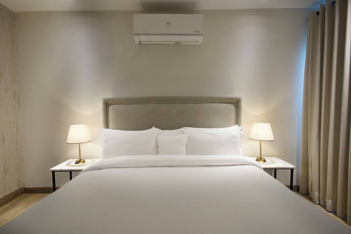 Une chambre d’hôtel avec un grand lit, deux tables de chevet avec des lampes, un climatiseur au plafond et une fenêtre avec des rideaux.