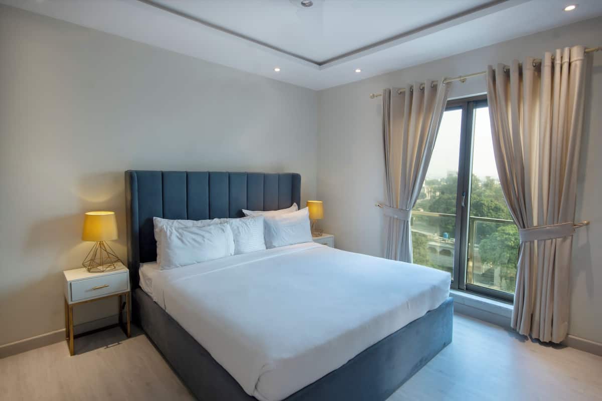 Une chambre moderne avec un grand lit, deux tables de chevet avec des lampes et une vue sur la verdure par la fenêtre.