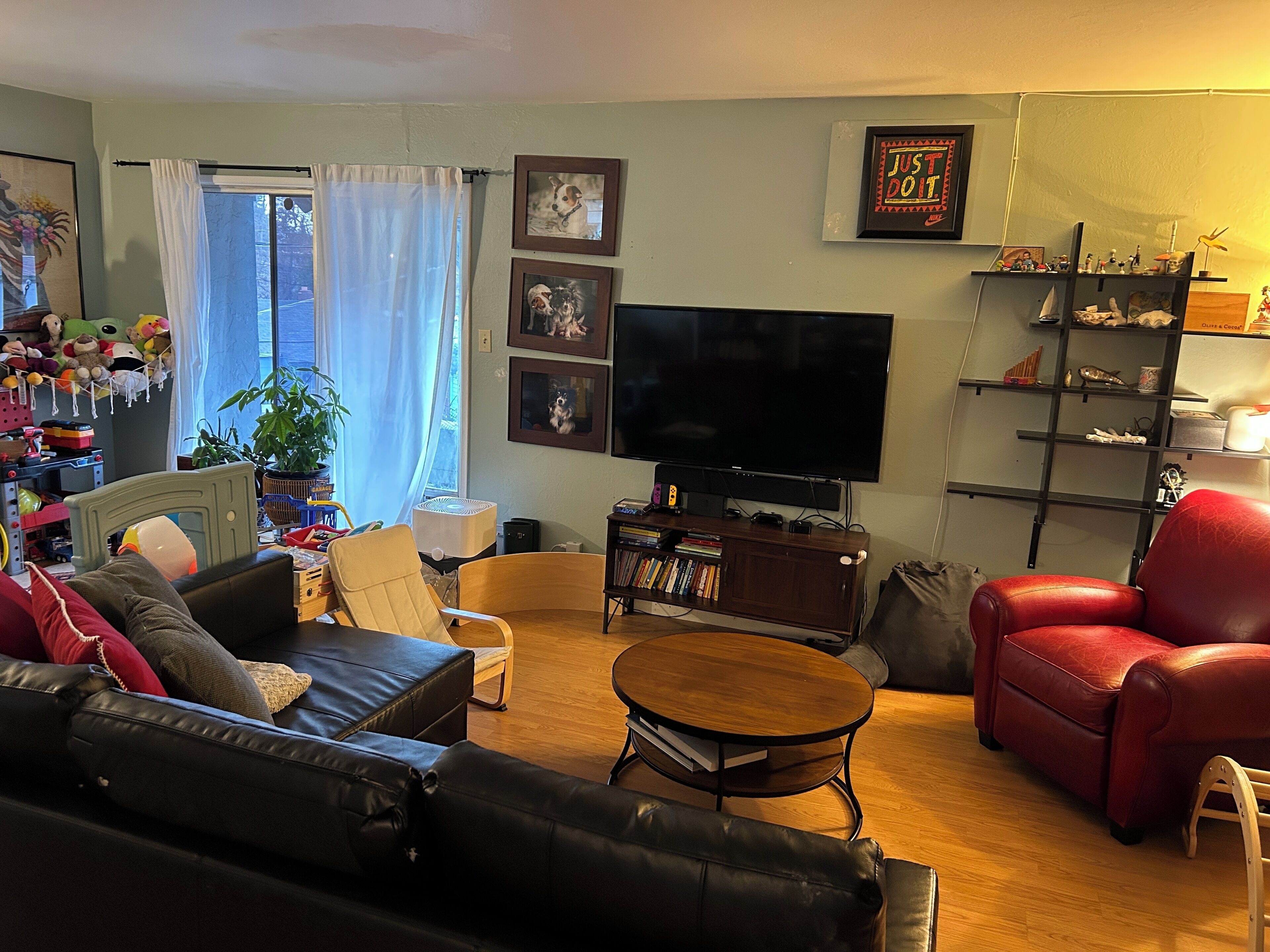 A living room with a black leather sofa, a red leather armchair, a wooden coffee table, and a television. There are framed pictures on the wall, a bookshelf, and a window with curtains.