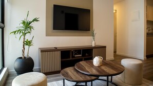 Una sala de estar moderna con un televisor de pantalla plana, una planta grande en maceta, una mesa de centro redonda de madera y un espacio de descanso acogedor.