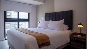 Une chambre d’hôtel moderne avec un grand lit, une tête de lit sombre, du linge de lit blanc, un plaid marron, une lampe de chevet et une vue sur la ville par une grande fenêtre.