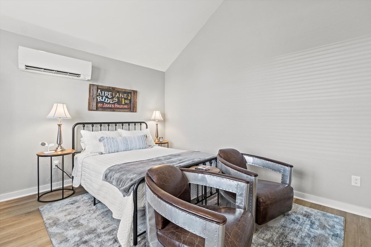 A bedroom with a bed, two bedside tables, a wall-mounted air conditioner, and two leather chairs.