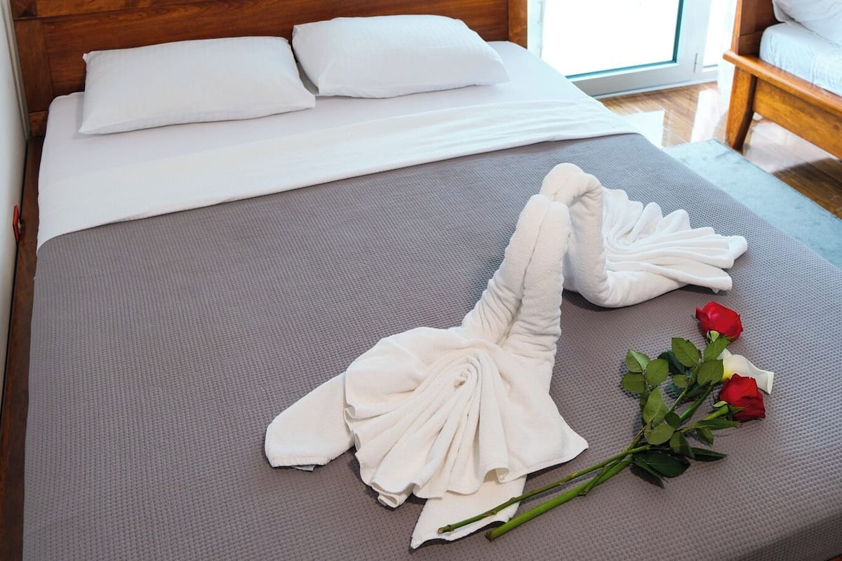 A neatly made bed with white linens and a grey mattress, a wooden headboard, and a wooden nightstand with a lamp.