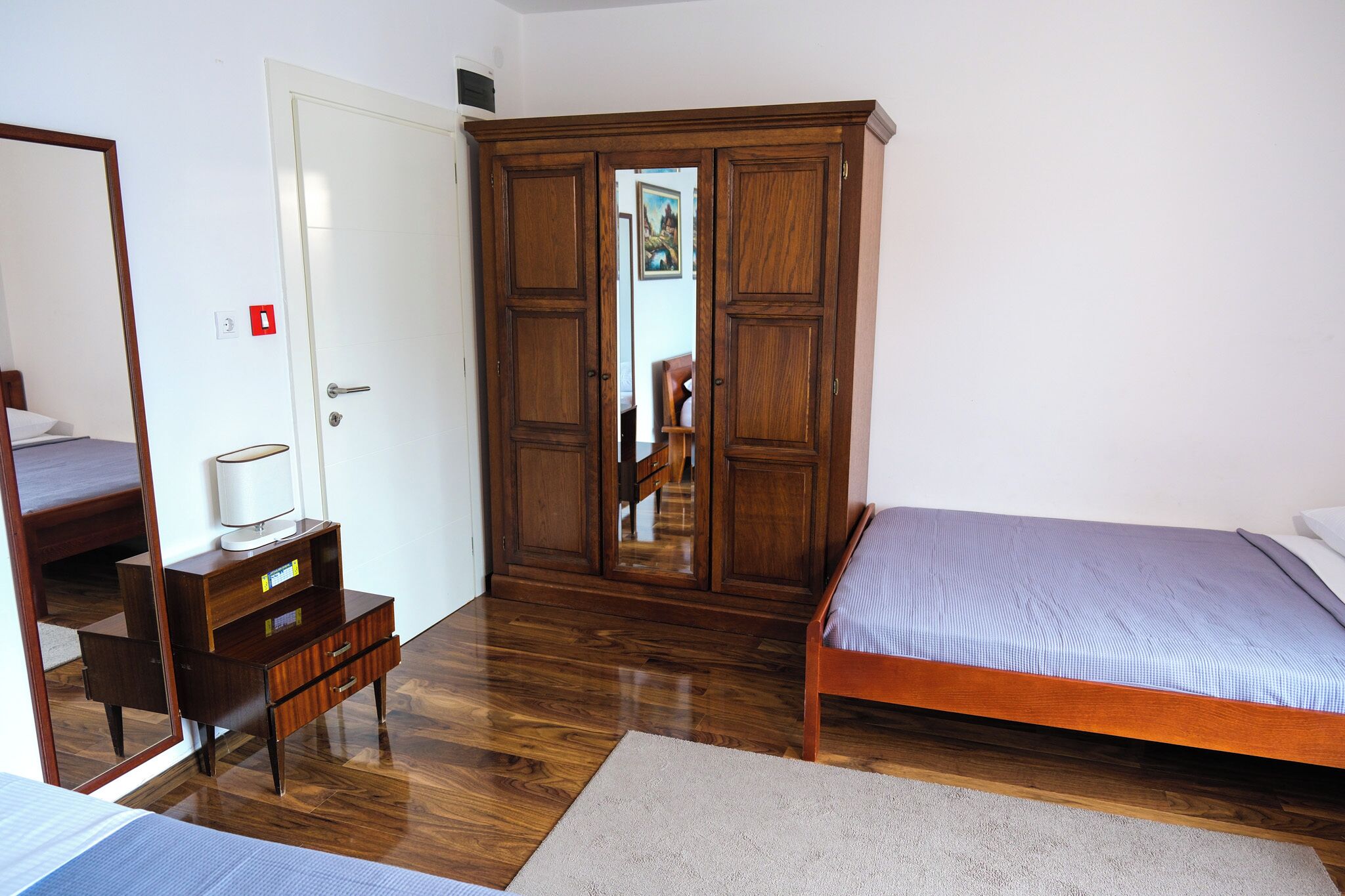 A bedroom with a wooden wardrobe, a bed, a bedside table with a lamp, and a mirror.
