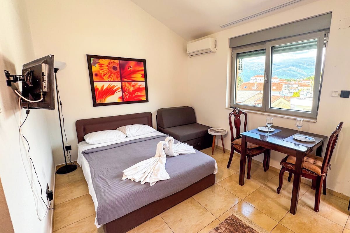 A hotel room with a bed, a sofa, a dining table with chairs, a TV, and a window with a city view.