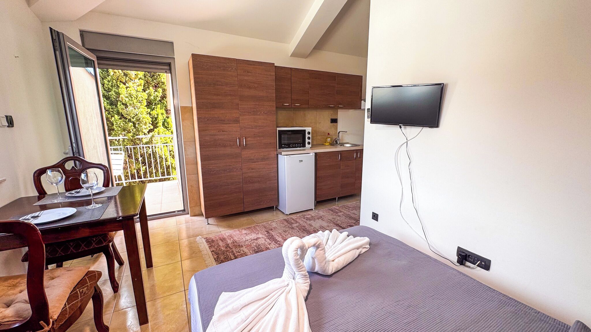 A compact room with a bed, a dining table with chairs, a kitchenette, and a TV mounted on the wall.