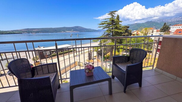 A balcony with two wicker chairs and a small table, overlooking a body of water and a mountainous landscape. - Qumi Apartments (Tivat)